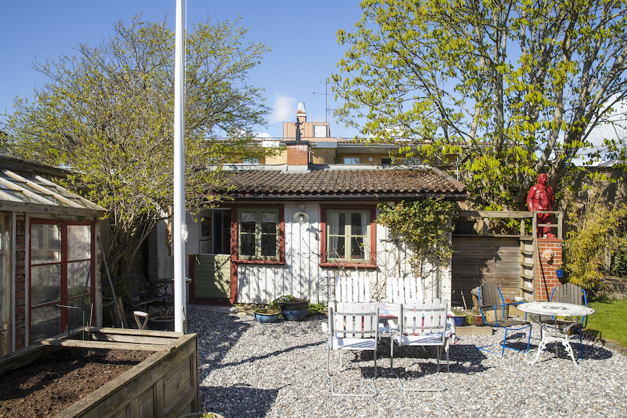 Stugan utvändigt med växthus och flaggstång till vänster, sittgrupp på grusad uteplats framför stugan.