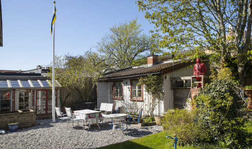 Stugan sedd från trädgården med växthus och flaggstång till vänster, sittgrupp på grusad uteplats framför stugan.