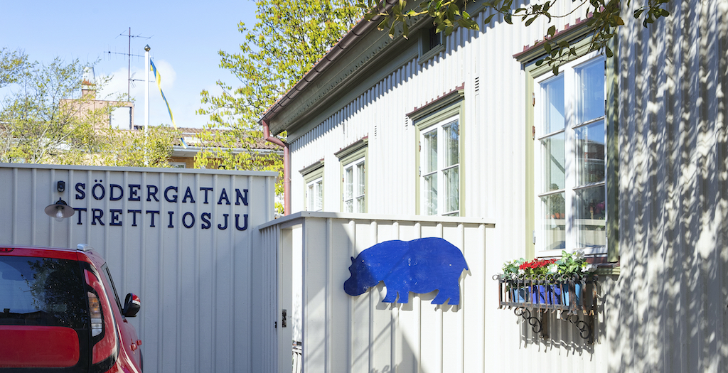 Entré till Södergatan 37. Ett gråvitt plank med gatunummer och en blå skylt i form av en flodhäst. En husvägg med fyra fönster.